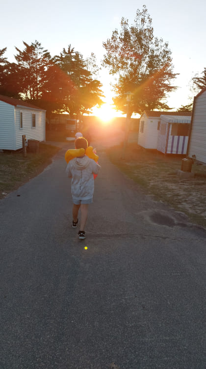 Une personne marche sur un chemin entre des mobile homes au coucher du soleil, portant un sweat à capuche et des tongs, avec un canard gonflable sur les épaules.