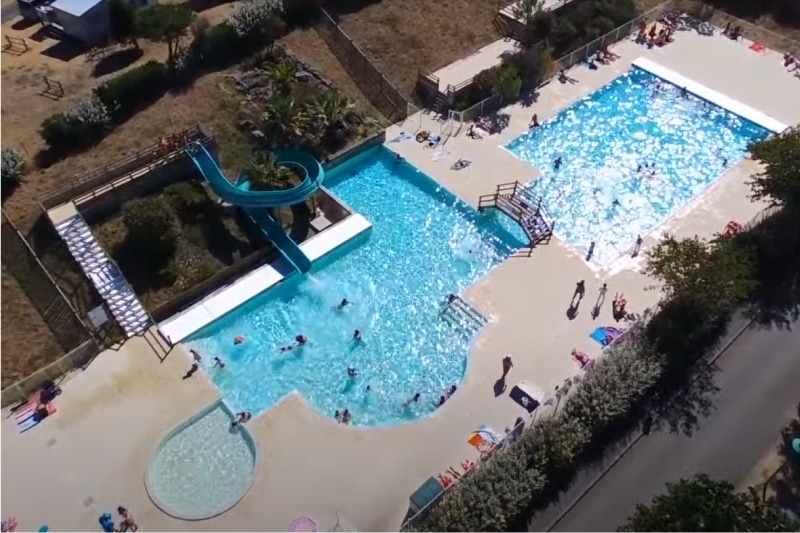 La photo montre une vue aérienne de deux piscines extérieures remplies de baigneurs et un toboggan aquatique bleu qui se termine dans l'une des piscines.