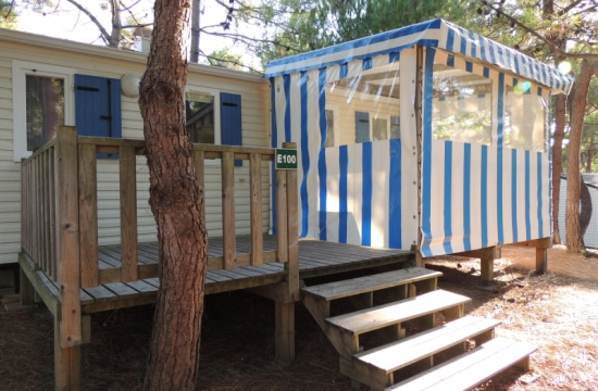 Une cabane en bois surélevée avec une terrasse et une tonnelle rayée bleu et blanc se trouve entourée d'arbres dans ce qui semble être un environnement de camping.